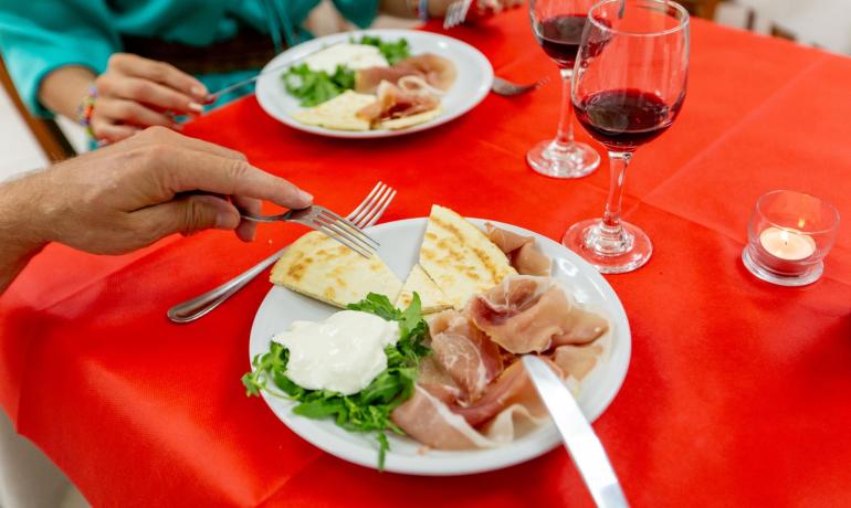 Piatto con prosciutto, formaggio, rucola e vino rosso su tavola rossa.