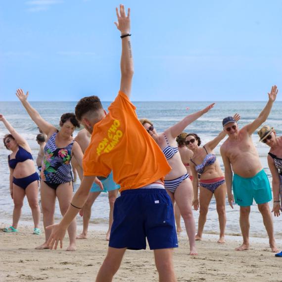 Des gens font des exercices sur la plage, sourires et mouvements sous le ciel bleu.