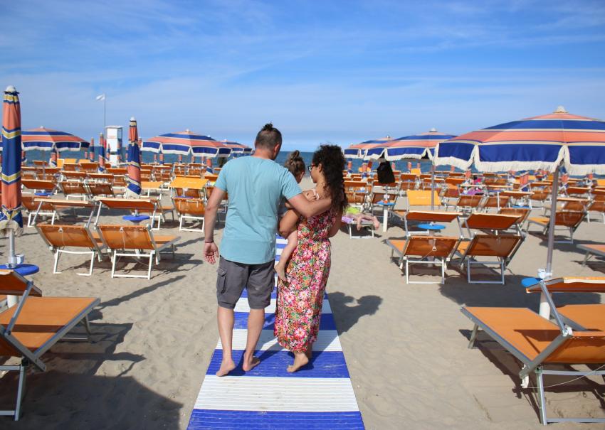 Famille marche sur la plage, soleil, parasols colorés et mer à l'horizon.